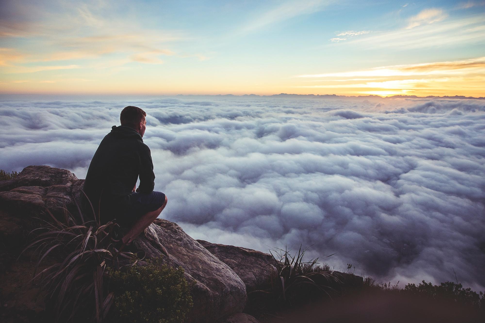 Man wearing beanie and jacket sitting on a top of a rock with a view full of clouds
