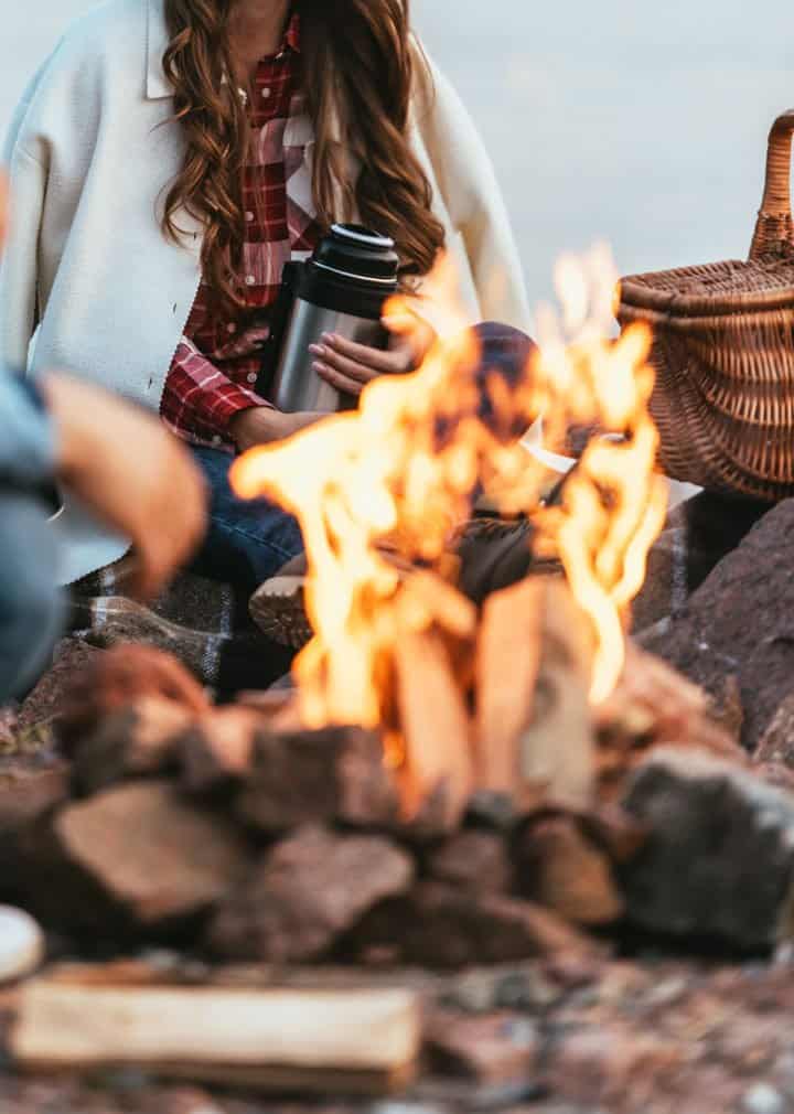 Lady sitting in front of bonfire with picnic basket holding a thermos. 