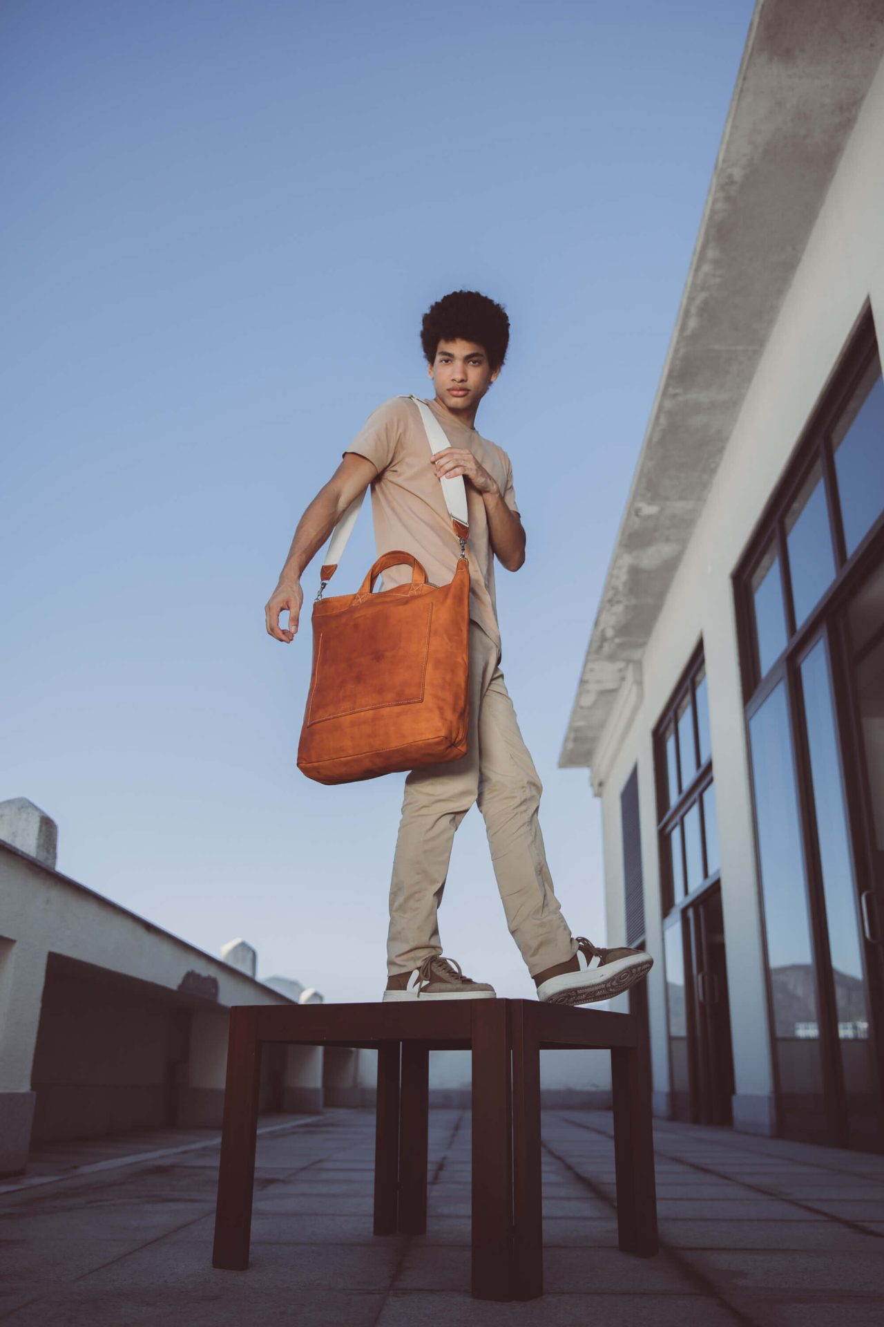 Handmade vegetable tanned leather carry on bag in cognac worn by male model in Rio de Janeiro over the shoulder with the off white webbed cotton adjustable strap.jpg
