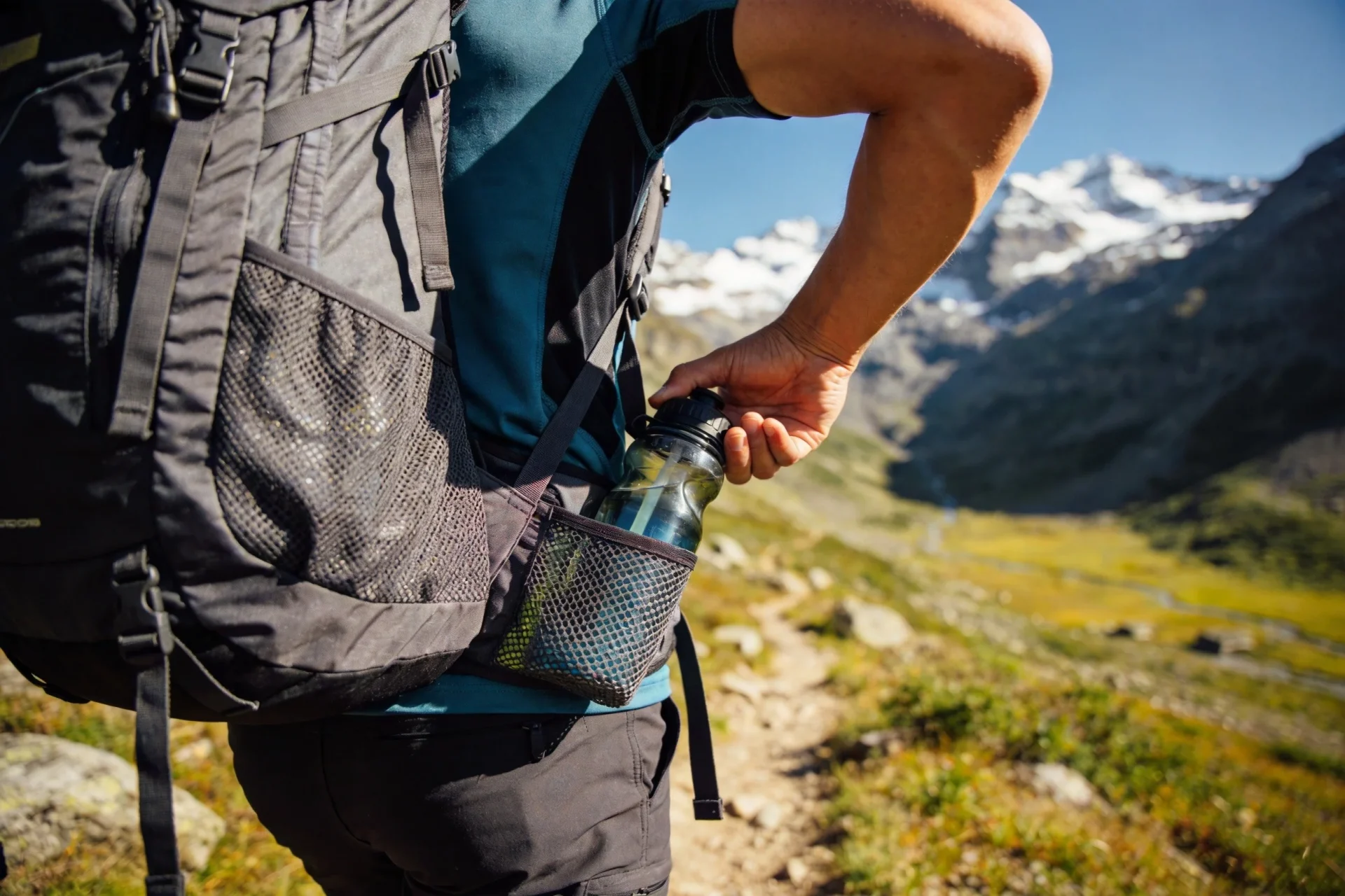 Athletic hiker on mountain trail reaching for water bottle from side mesh pocket of hiking backpack with alpine landscape